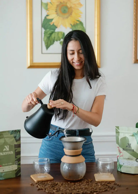 Woman Owner of Nativa pouring coffee from a black kettle into a Chemex Pour Over on a wooden table.