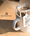 White mug with coffee filter and coffee bags, next to a box labeled 'Drip Coffee Bags'.